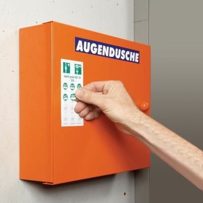 Eine Geprueft -Plakette wird auf ein Jahres -Grundetikett mit zwoelf Feldern, zwei Piktogrammen für Augen- und Körperdusche geklebt. Die Überschrift des Grundetikettes ist: Geprueft gemaeß BGR 120 und der Jahreszahl